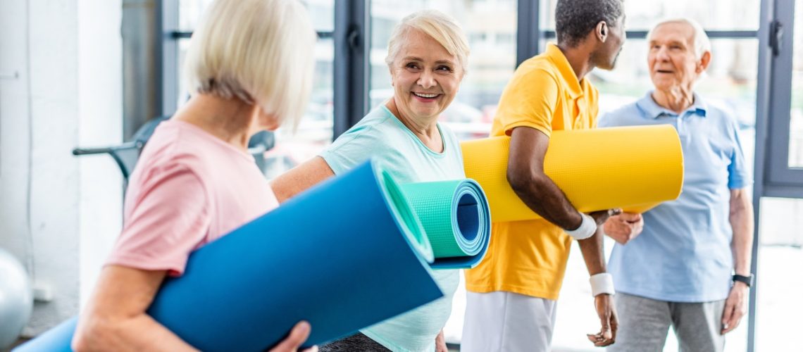 senior sportswomen holding fitness mats and their male friends standing behind at gym